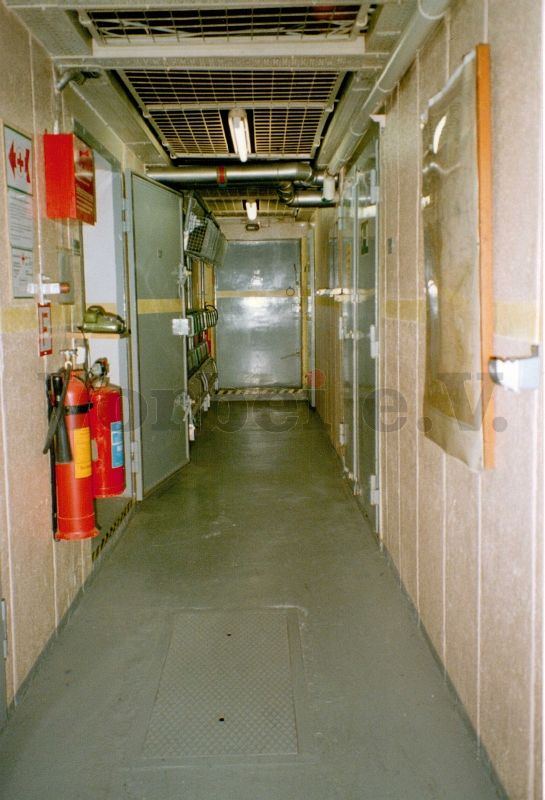 Raum 45 (Vorraum der Maschinenräume): Kleinlöschgeräte in Wandhalterungen und Fernsprechtischapparat auf einem Wandpult. Die geöffnete Tür führt in Raum 24 (Lüftungs- und Klimaanlage). Auf der rechten Seite befindet sich die geschlossene Tür zum Raum 20 (Netzersatzanlage und Gleichrichter).