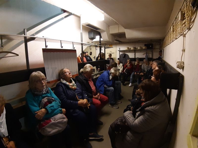 Viele Personen sitzen auf den Schutzraumbänken im Bunker. Über die originale Rundsprechanlage wird die Kurzgeschichte "Krieg spielen - Frieden lernen" von Herrn Gerd Zietlow vorgelesen.