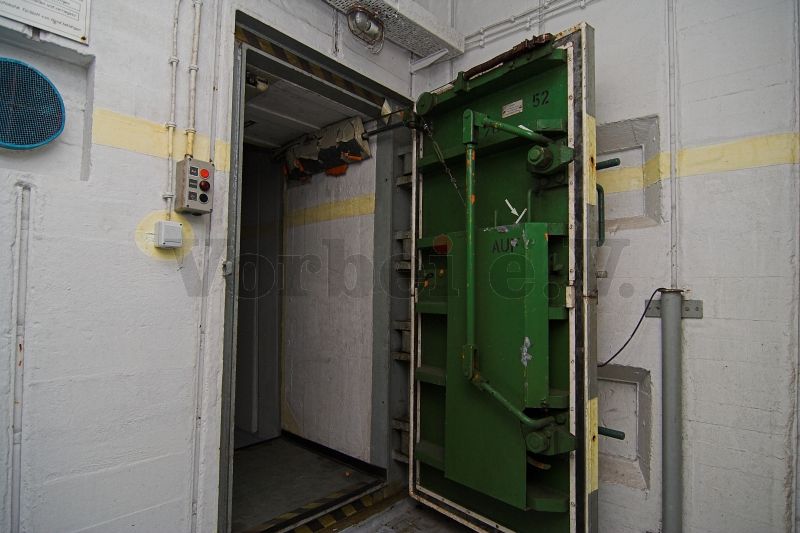 Innere Panzerdrucktür in der Zugangsschleuse (Raum 52). Die Zusammenfassung der Türenhälften sowie das Hebelgestänge zur kraftschlüssigen Verbindung der beiden Vorreiber ist deutlich erkennbar. Links neben der Tür befinden sich die Drucktaster für die Bedienung der Anlage.