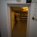 Blick aus einem Liegeraum durch die geöffnete Tür in den Gangbereich des Bunkers. Im Gangbereich entlang befinden sich Schutzraum-Sitzplätze.