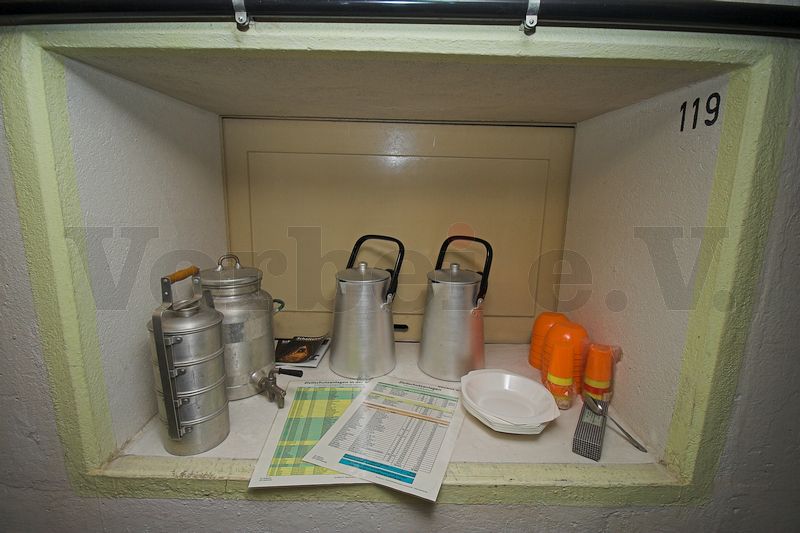 Die Getränkekanne hat einen Platz im Bunker in der Durchreiche der Küche, die zur Essensausgabe dient, bekommen. In der Durchreiche sind bereits eine weitere identische Kanne, Getränkebecher, Essschalen und Kunststoffteller, eine Getränkekanne mit Zapfhahn sowie ein vierteiliger Essensbehälter aufgestellt.