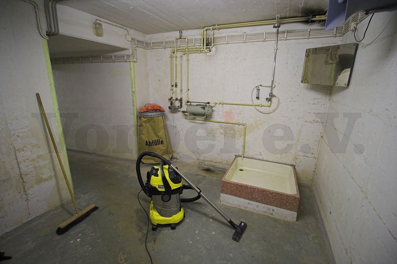 Ein Schleusenbereich im Museumsbunker Hannover wird gereinigt. Auf dem Boden steht ein Mehrzwecksauger. An der Wand lehnt ein Besen. In der Schleuse befindet sich eine Dusche zur Dekontamination.