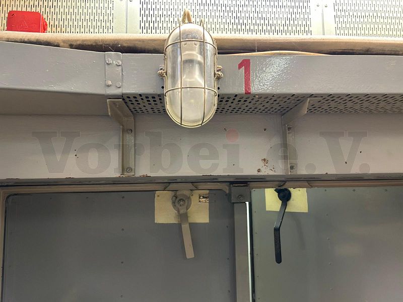Im Raum 20 (Netzersatzanlage und Gleichrichter) wurde die Notbeleuchtungslampe senkrecht an einer Kabelbühne montiert, die oberhalb der Zugangstür verläuft. Hier wurde die rote „1“ rechts neben der Lampe direkt auf der Kabelbühne angebracht.