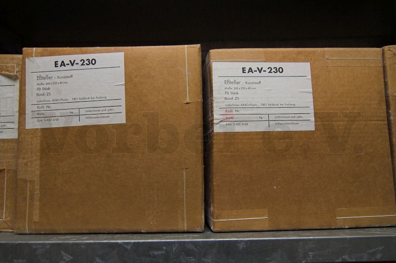 Verschlossene Kartons in einem Regal. Die Kartons sind beschriftet mit Eßteller Kunststoff, Maße 260 x 232x 40 mm, 70 Stück, Bund ZS.