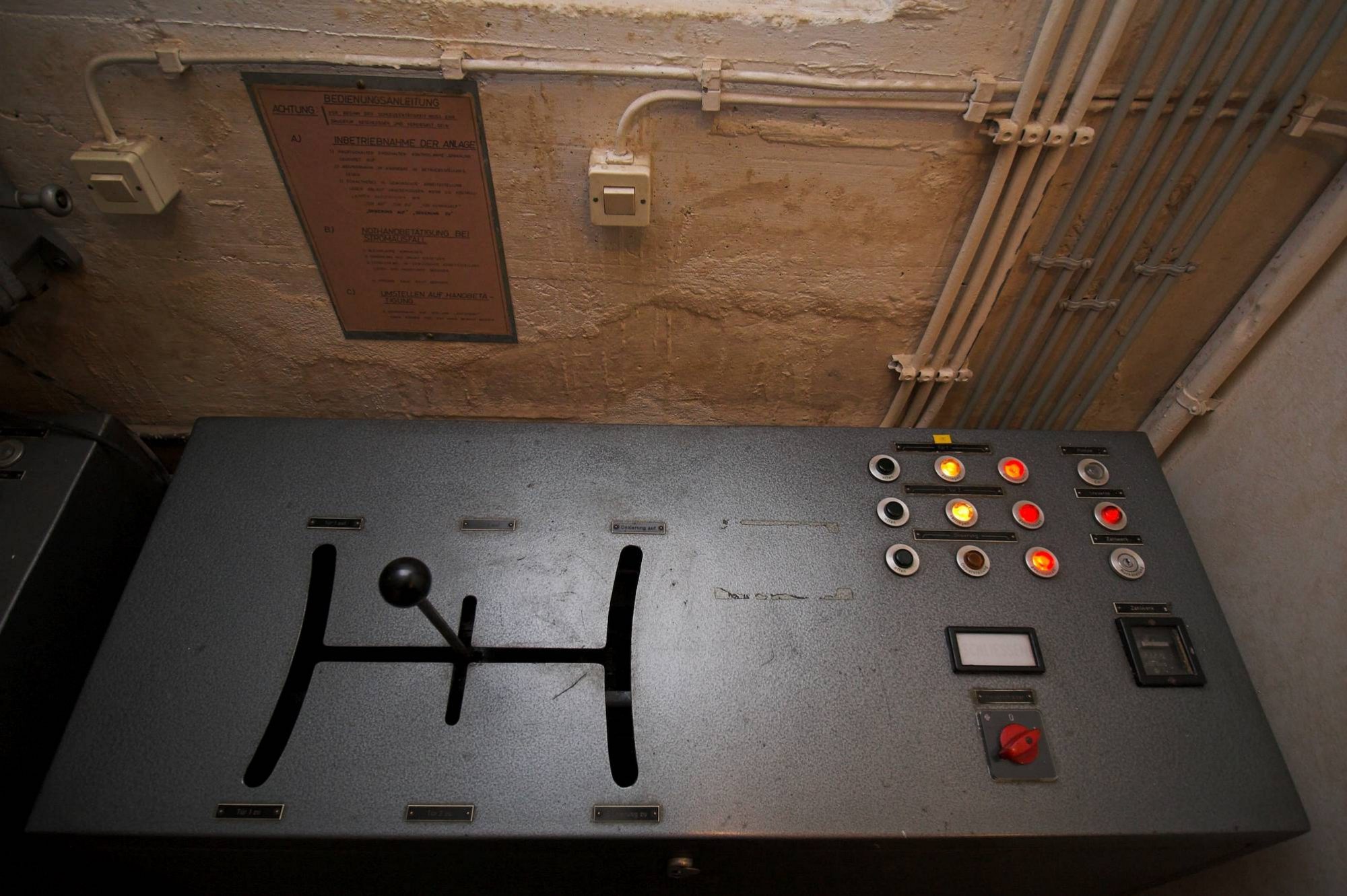 Ein Steuergerät für eine einzelne Schleusenanlage im Museumsbunker Hannover: Mit dem Schalthebel im linken Bereich kann jeweils die innere und die äußere Schleusendrucktür sowie die Dosierschleuse auf- und zugefahren werden. Die Kontrolllampen signalisieren, dass alle Türen der Schleusenanlage geschlossen und verriegelt sind. Das Steuergerät enthält alle technischen Komponenten, die für den Betrieb der elektrohydraulischen Schleusentüren benötigt werden.