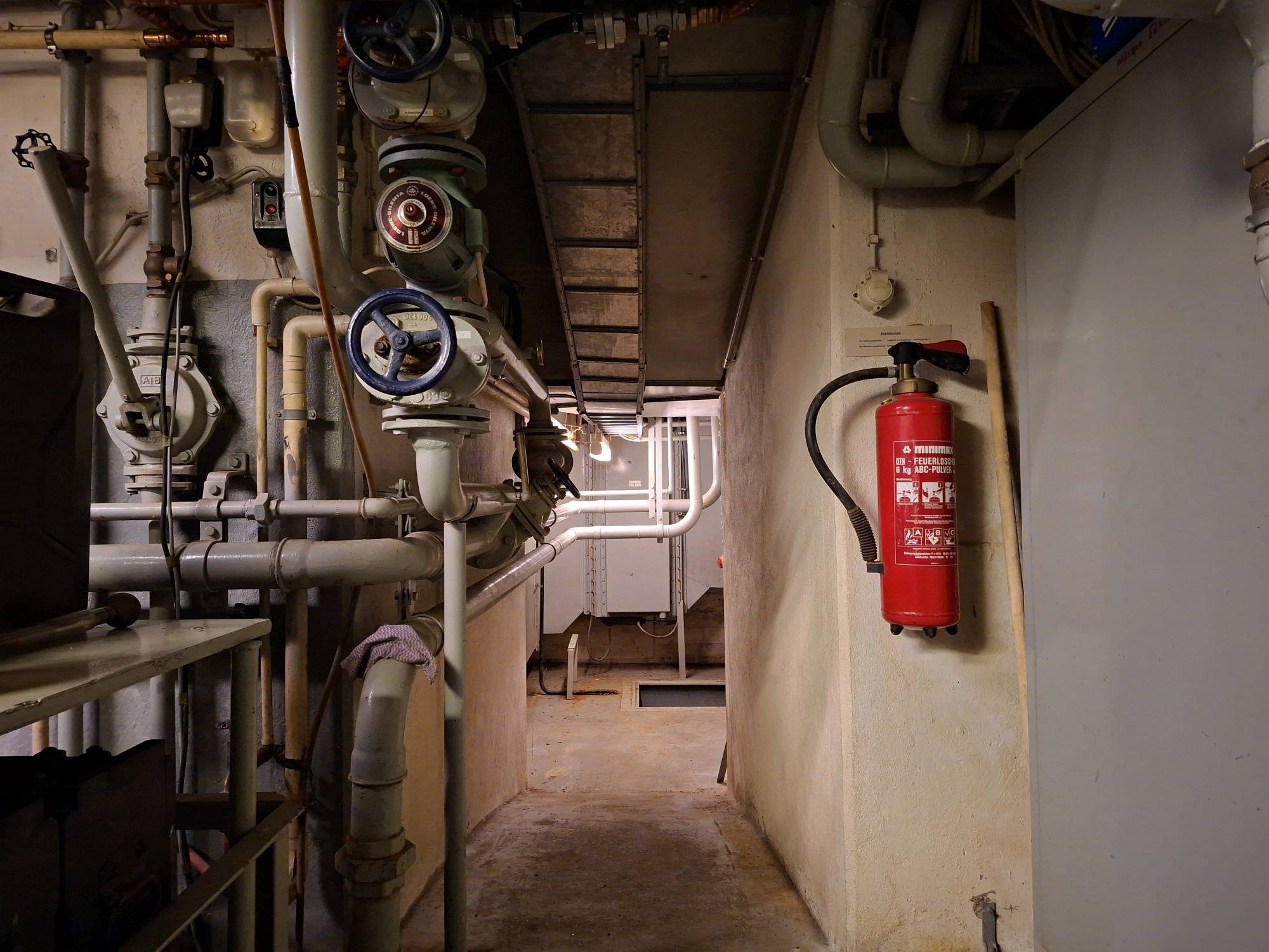 Durchgang zum Abluftbereich des Filteranbaus im Museumsbunker Hannover. Die beiden Rohrleitungen in der Bildmitte bilden den Vor- und Rücklauf der Kühlwasserleitung zu den Wärmetauschern, die sich im 1. Obergeschoss des Filterbauwerkes befinden. Dahinter befindet sich der Abluftkanal mit der elektropneumatisch betätigten Abluftklappe.